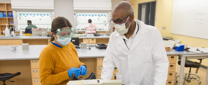Science student and professor wearing masks and goggles in lab.